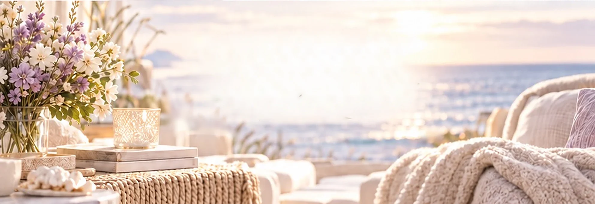 A seaside room at sunset: lavender and white flowers, a glowing candle on stacked books, the ocean beyond, and a bed draped in a soft knit throw.